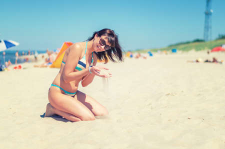 Young beautiful girl in bikini and sunglasses sit on sandy beach, pour sand through fingers and smile, Baltic sea water, tourist female on vacation in Lithuania Curonian Spit in sunny summer dayの写真素材