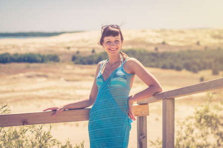 Young beautiful girl in bikini, pareo wrap and sunglasses on head posing near wooden fence, smile and looking at camera, tourist female on vacation in dune of Lithuania Curonian Spit in summer dayの写真素材