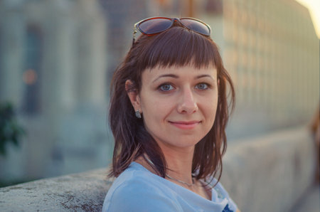 Close-up portrait of young cheerful beautiful woman with dark hair, gray eye and sunglasses on head, looking at camera, posing and smiling outdoors in city, blurred backgroundの写真素材