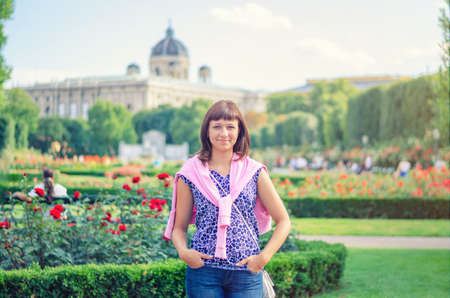 Young beautiful caucasian girl with flowered shirt looking at camera, posing and smile in Volksgarten garden park with flowers in Vienna city historical center in sunny summer dayの写真素材