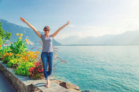 Young beautiful girl traveler with striped shirt and jeans posing, raise hands and smile on embankment of Lake Leman Geneva in Montreux town of Swiss Riviera, mountains Alps background, Switzerlandの写真素材