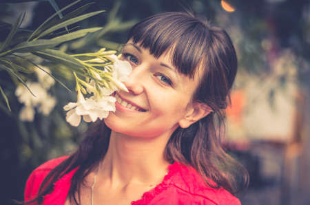 Close-up portrait of young beautiful caucasian girl with red jacket looking at camera, smile and smell white flowers with blurred background, woman with dark hair and gray eyeの写真素材
