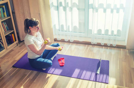Young beautiful woman in sportswear doing fitness workout at home, exercising raising dumbbells biceps curl, sitting on floor violet mat crossing legs and looking at tablet screen in living roomの写真素材