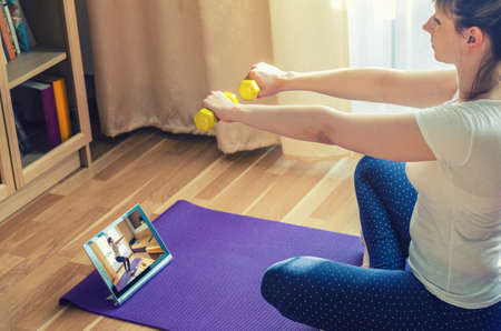 Young woman doing workout sports at home, raising dumbbells with straight arms, sitting on floor mat and watching online tutorials video on tablet screen in living room, sport and recreation conceptの写真素材