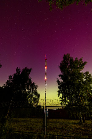 A tall communication tower under a starry night sky with purple hues, surrounded by trees and a fence.の写真素材