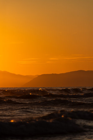 Golden sunset over ocean waves with distant mountains.の写真素材