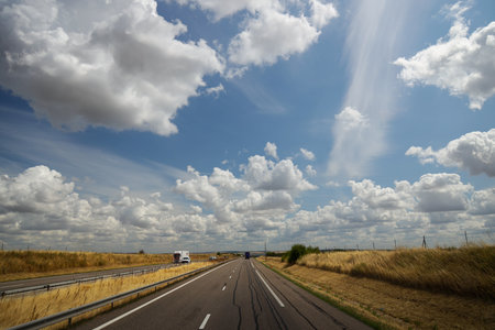 A scenic view of a highway stretching into the distance under a blue sky filled with fluffy clouds, surrounded by golden fieldsの写真素材