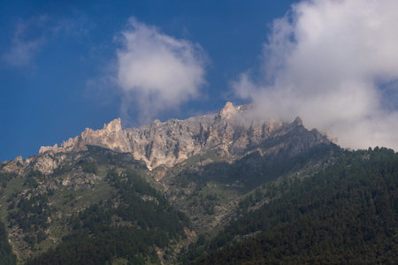 Mountain peak with clouds and forested slopes under a blue sky.の写真素材
