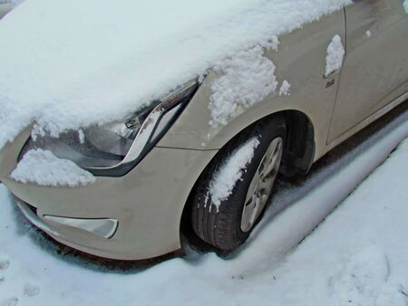 Front headlight of a passenger car and the left wheel, photographed in close-up, covered with snow after heavy snowfall, in a parking lot. Vehicles in the winter.の写真素材