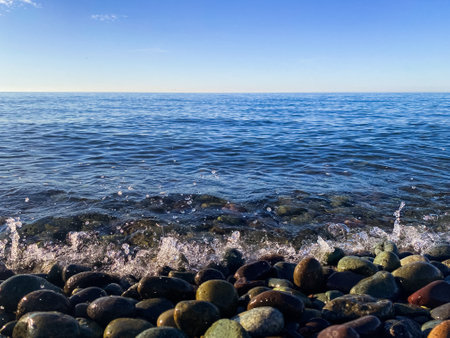 Close-up of ocean waves splashing on smooth pebbles under a bright blue sky, creating a serene coastal scene.の写真素材