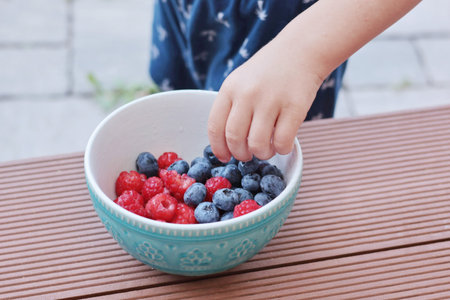 Child hand pick berries from bowl. Blueberries and raspberries. Family and lifestyle. Backyard in summer and nature, terrace. . High quality photoの写真素材