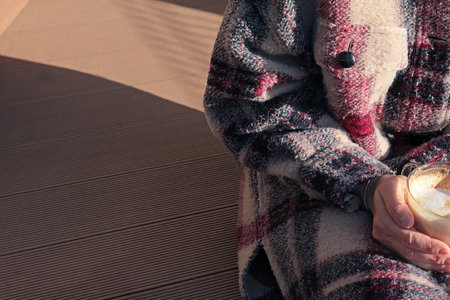 Elderly woman in warm fashion wool coat in fall on terrace with cup of cappuccino coffee on sunny...