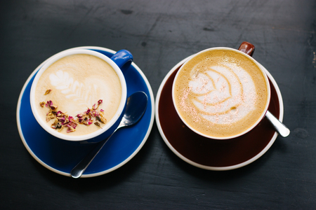 Two cups of cappuccino with latte art on black background. Beautiful foam, blue and brown ceramic cups, stylish toning, place for design and text.の写真素材