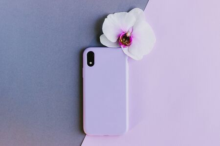 Lilac silicone case on grey and pink background. Pastel purple silicone case for smartphone. Protection for the back cover of the mobile phone with a slot for the camera. Orchid flower.の写真素材
