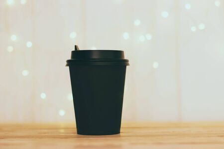 Black coffee Cup on a wooden table. A black Cup with a lid against a blurred wooden background with warm bokeh bulbs. Cafe. Selective focus.の写真素材