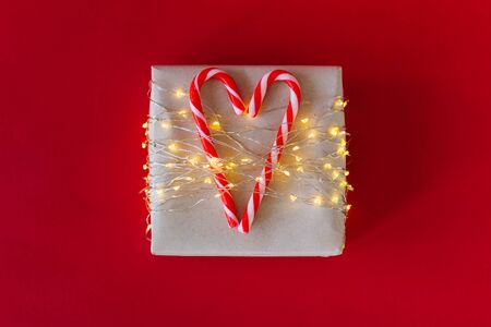Gift box on bright red background. Christmas striped candy canes. The gift is tied with led tape. Two lollipops are connected in the heart. Candy heart. Christmas garland. Selective focus.の写真素材