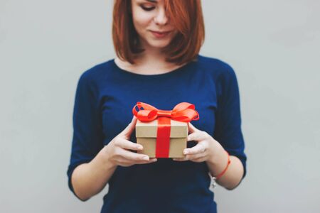 A girl with short red hair in a blue jumper holds a gift tied with a red satin ribbon. Portrait of a girl with a gift box in her hands. Gift for Christmas, birthday, Valentines day. Selective focus.の写真素材