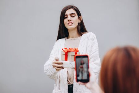 The girl is photographed on a smartphone. A girl in a white knitted blouse with gifts in her hands is photographed on a smartphone. Girl happy holding a gift box with a bow. Selective focus.の写真素材