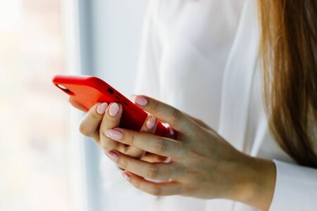 Red smartphone in a woman's hand close-up. The girl holds the phone in a red plastic case. Neat gentle light manicure. Selective focus.の写真素材