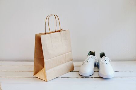 Fashionable white sneakers and a pack of brown environmental paper on a light background. Paper bag on light wooden floor. Simple brown paper shopping bags. Environmental ship packages. Sports shoes.の写真素材