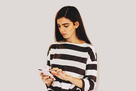 A girl in a striped sweater holds a smartphone in her hands. The girl is typing a message on her phone. Girl on a gray background. Selective focus.の写真素材