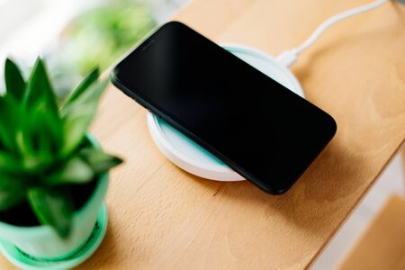 Smartphone in mint silicone case is charged from a wireless charger. The mobile phone is charged on a wooden nightstand or shelf.の写真素材