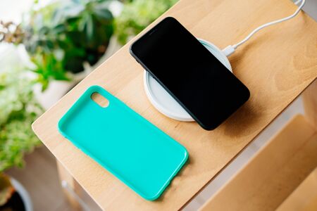Black smartphone and mint silicone case is charged from a wireless charger. The mobile phone is charged on a wooden nightstand or shelf.の写真素材