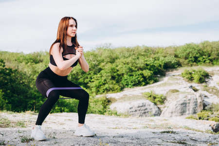 A beautiful athlete in the mountains. The girl is engaged in sports in nature. Purple rubber band for stretching and swaping muscles of hands and feet.の写真素材