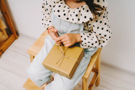 A little girl in a denim jumpsuit sits on a wooden chair and holds a gift box on her knees.の写真素材