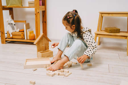 A girl with two ponytails on her head sits on the floor in her room and plays with wooden cubes. The girl sits on the floor and builds a tower out of the constructor.の写真素材