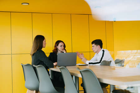 A group of people in the conference room with bright yellow walls sit behind a laptop and discuss the project. The guy's writing in a notebook.の写真素材