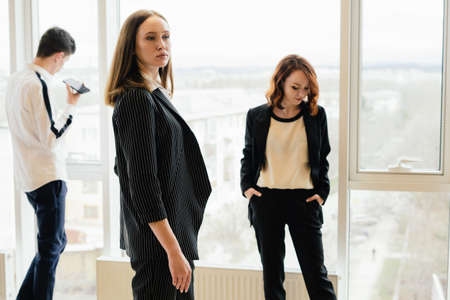 A group of three young people stands at the panoramic window of their company. Beautiful girls and a...