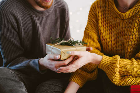 A girl and a guy in gray and yellow sweaters exchange a gift. Gift craft box with a coniferous branch in the hands of a girl and a guy.の写真素材
