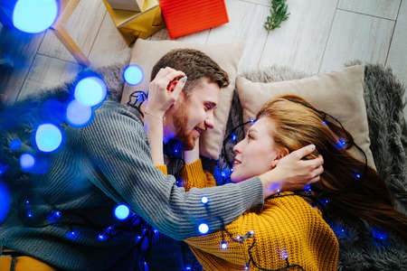 Beautiful couple in love lies on the floor and hugs. Blue bokeh. A happy guy and a girl lie on a soft gray blanket. Christmas blue garland.の写真素材