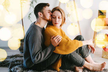The guy gently kisses and hugs his girlfriend. Romantic couple of lovers against the background of a Christmas tree made of natural wood. Yellow bokeh.の写真素材