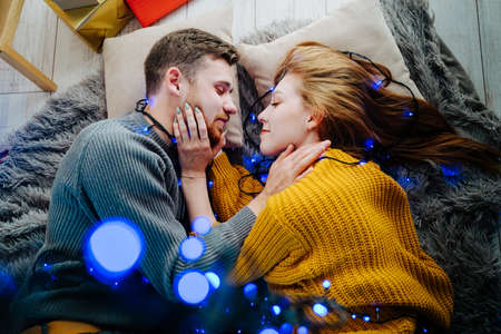 Beautiful couple in love lies on the floor and hugs. Blue bokeh. A happy guy and a girl lie on a soft gray blanket. Christmas blue garland.の写真素材