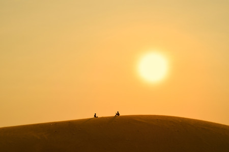 Silhouettes of people sitting and chatting at the sandy hill under golden sunset sunの写真素材