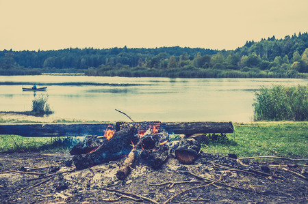 Rustic bonfire by the lake in the forest on the background of blurry boat with fisherman on the lake, vintage photo.の写真素材