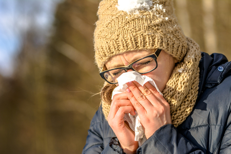 Sneezing woman blowing her nose with a tissue. Sick woman with cold and flu in winter, outdoors. Girl in warm clothes - hat and scarfの写真素材