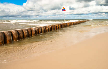 Landscape of beach and sea with waves and cloudy sky. Breakwater and warning sign in the foreground. Baltic sea coast near Leba in Poland.の写真素材