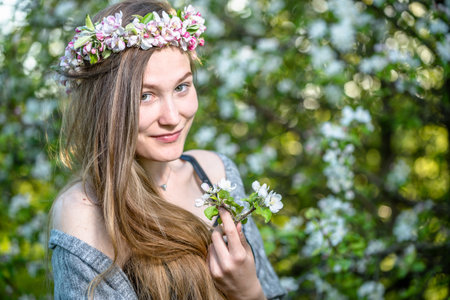 Cute girl with a flower crown on head posing with a flowering branch on a blooming tree background.の写真素材