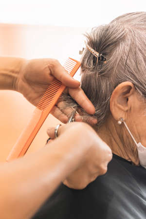 Medium short shot of female stylist trimming with scissors and orange comb the wet hair of an elderly client in a hairdressing salon on blurred background. Working woman, lifestyle. Older client complying with hygiene measures.の写真素材
