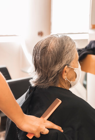 Medium short shot of female stylist preparing seat to cut wet hair of elderly client in a hairdressing salon on blurred background. Working woman, lifestyle. Older client complying with hygiene measures.の写真素材