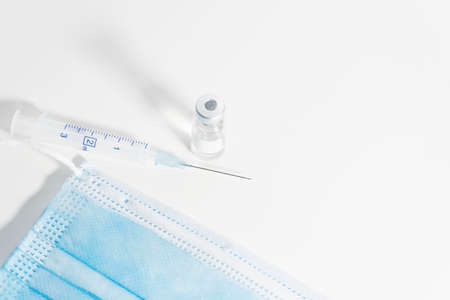 Vaccination concept. COVIC-19 coronavirus vaccine. White table with blue surgical mask, vaccine vial bottle and syringe. White background.の写真素材