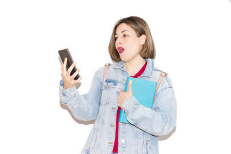 Young caucasian female student isolated on a white background surprised by the video she is watching on her mobile phone, while holding a book with the other arm. Lifestyle, spring.の写真素材