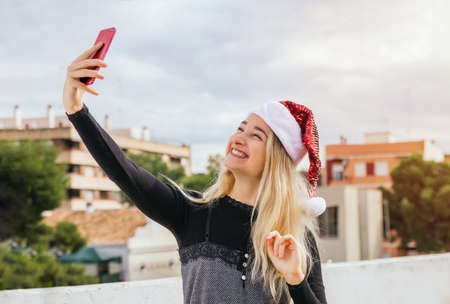 Stock photo of a beautiful blonde woman taking a selfie with mobile phone for Christmas day. She is laughing and having fun. Xmas vlogging, technology, social networks and people conceptsの写真素材