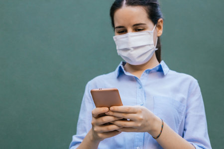 Stock photo of a young woman wearing a face mask chatting with her phone on a green background. Selective focusの写真素材