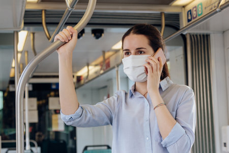 Stock photo of a young woman wearing a face mask talking on the phone while standing on the tramの写真素材