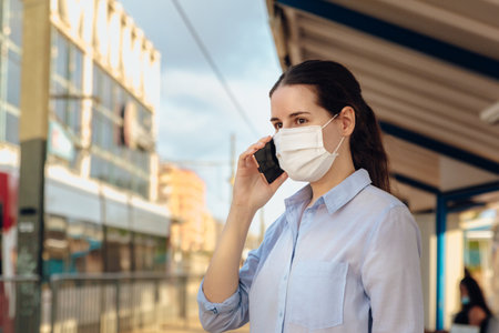 Stock photo of a young woman wearing a face mask and talking on the phone while waiting for public transport. New normalの写真素材