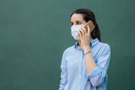 Stock photo of a young woman wearing a face mask talking on phone on a green background and copy space on the leftの写真素材